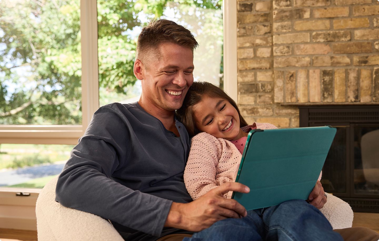 Father and daughter sitting together on a chair, smiling and looking at a tablet in a cozy living room with a large window and brick fireplace.